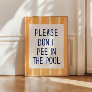 May include: A framed art print with the text "PLEASE DON'T PEE IN THE POOL" in navy blue. The artwork has a white background with the text, set against a yellow and orange striped background. The frame is a light wood color.