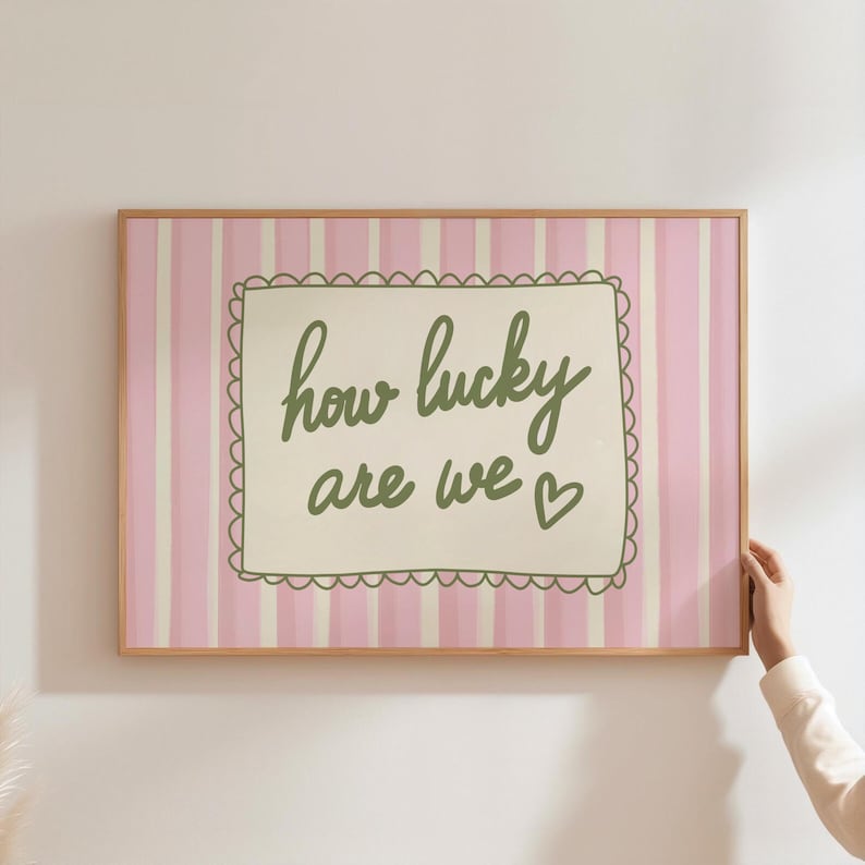 May include: A framed print with a pink and white striped background. The print features the phrase "how lucky are we" in green script within a scalloped-edged rectangle. A small heart is at the end of the phrase.