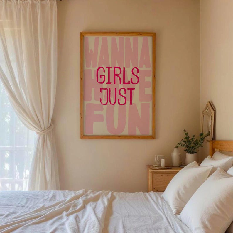 May include: A rectangular wall art print in a light wooden frame featuring the text "GIRLS JUST WANNA HAVE FUN" in bold, dark pink lettering against a pale pink background, displayed on a plain wall above a bed with white bedding.