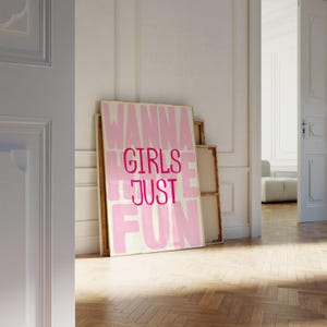 May include: A framed canvas print with the words "WANNA GIRLS JUST FUN" in pink and white. The artwork leans against a white wall in a room with a wooden floor. Additional canvases are stacked behind it.