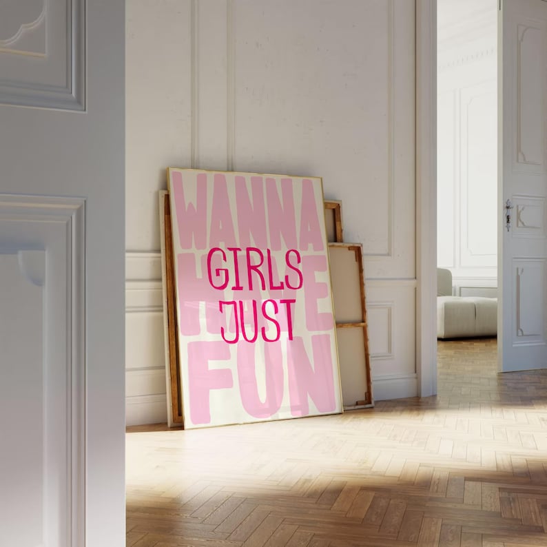May include: A framed canvas print with the words "WANNA GIRLS JUST FUN" in pink and white. The artwork leans against a white wall in a room with a wooden floor. Additional canvases are stacked behind it.