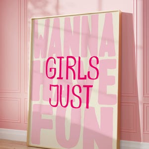 May include: A framed art print with the words "WANNA GIRLS JUST FUN" in pink lettering on a cream background. The print is in a gold frame and leans against a pink wall.