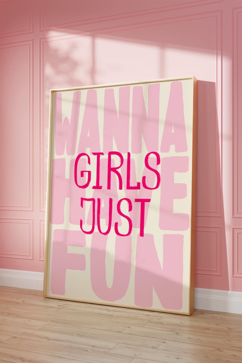 May include: A framed art print with the words "WANNA GIRLS JUST FUN" in pink lettering on a cream background. The print is in a gold frame and leans against a pink wall.