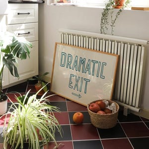 May include: A framed sign with the text "Dramatic Exit" in teal blue lettering with an arrow pointing to the right. The sign is hanging on a white wall next to a white radiator. There are plants and a basket of apples in the foreground.
