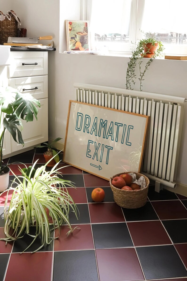 May include: A framed sign with the text "Dramatic Exit" in teal blue lettering with an arrow pointing to the right. The sign is hanging on a white wall next to a white radiator. There are plants and a basket of apples in the foreground.