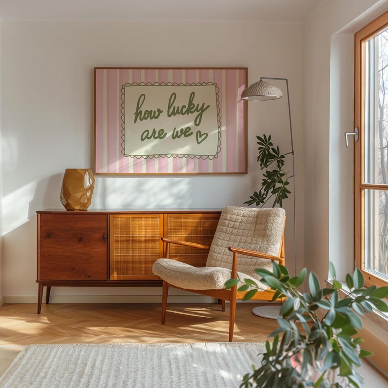 May include: A mid-century modern living room with a wooden cabinet, a beige armchair, and a framed artwork that reads "how lucky are we". A gold vase and a floor lamp with a woven shade complete the decor.