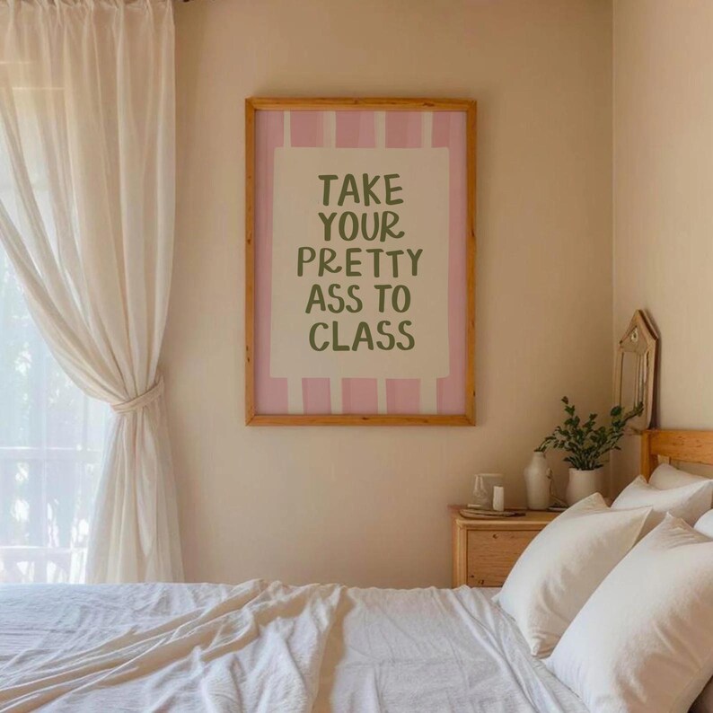 May include: A framed print with the text "TAKE YOUR PRETTY ASS TO CLASS" in a bedroom setting. The print has a pink and white striped border and a light brown wooden frame. The room has a bed with white linens.