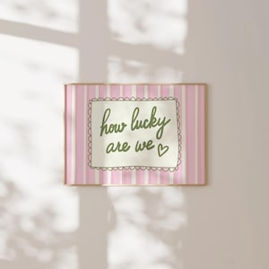 May include: A framed print with a pink and white striped background. The print features the phrase "how lucky are we" in a handwritten style, enclosed in a decorative border. A small heart is included at the end of the phrase.