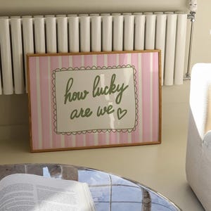 May include: A framed print with a pink and white striped background. The print features the phrase "how lucky are we" in green script within a decorative border. The frame is a light brown color.