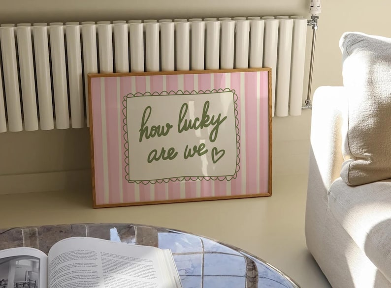 May include: A framed print with a pink and white striped background. The print features the phrase "how lucky are we" in green script within a decorative border. The frame is a light brown color.