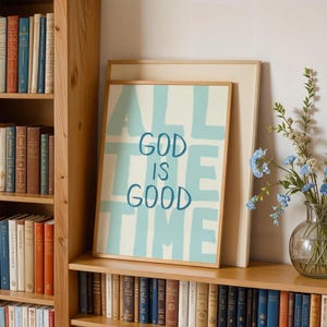 May include: A framed print with the words "ALL GOD IS GOOD TIME" in light blue on a cream background. The print is in a wooden frame and rests on a wooden shelf with a bookcase filled with books. A vase of flowers is to the right.