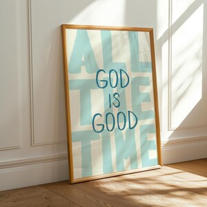 May include: A framed art print with the words "ALL THE TIME GOD IS GOOD" in blue lettering on a cream background. The wooden frame leans against a white wall and brown floor, with sunlight casting shadows.