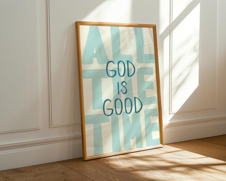 May include: A framed art print with the words "ALL THE TIME GOD IS GOOD" in blue lettering on a cream background. The wooden frame leans against a white wall and brown floor, with sunlight casting shadows.