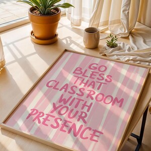 May include: A framed print with the text "GO BLESS THAT CLASSROOM WITH YOUR PRESENCE" in pink lettering on a pink and white striped background. The print is in a light wood frame and is displayed on a wooden surface.