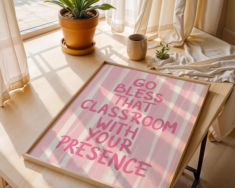 May include: A framed print with the text "GO BLESS THAT CLASSROOM WITH YOUR PRESENCE" in pink lettering on a pink and white striped background. The print is in a light wood frame and is displayed on a wooden surface.