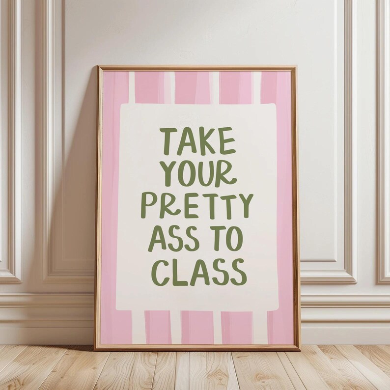 May include: A framed print with a pink and white striped background and the text "TAKE YOUR PRETTY ASS TO CLASS" in green lettering. The print is in a wooden frame and leans against a white wall.
