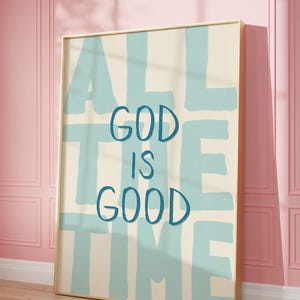May include: A framed art print with the words "ALL THE TIME GOD IS GOOD" in blue lettering on a cream background. The artwork is displayed against a pink wall and wooden floor.