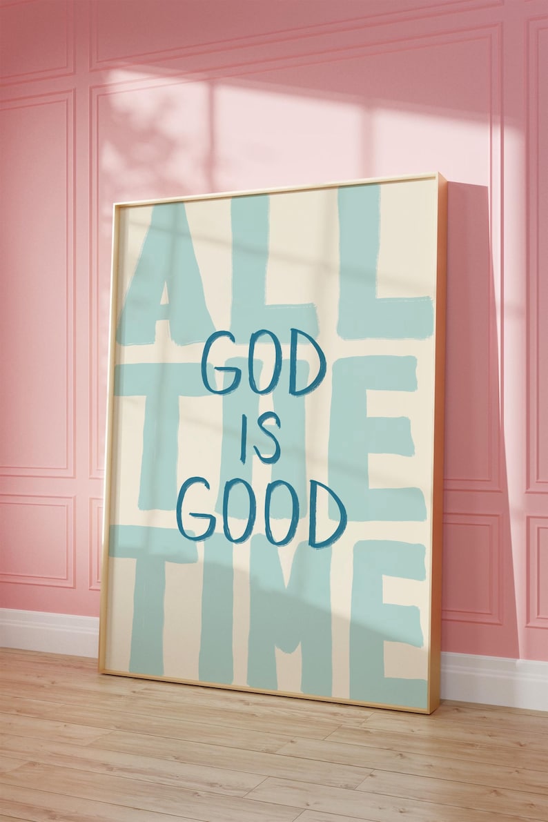 May include: A framed art print with the words "ALL THE TIME GOD IS GOOD" in blue lettering on a cream background. The artwork is displayed against a pink wall and wooden floor.