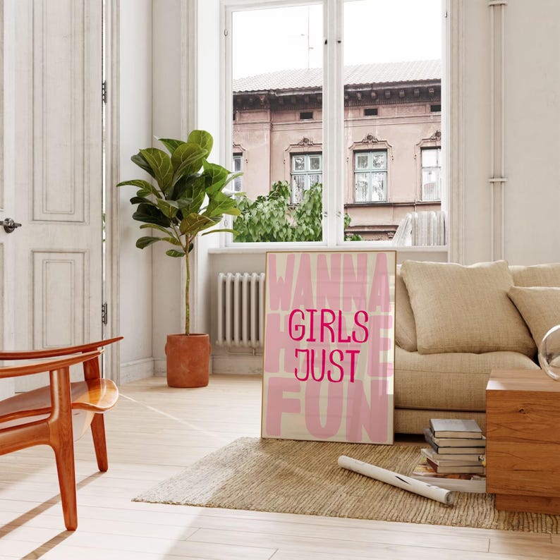 May include: A framed art print with the words "WANNA GIRLS JUST FUN" in pink and white. The print is displayed in a bright room with a beige sofa, wooden chair, and a potted plant. A window overlooks a building.