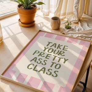 May include: A framed print with the text "TAKE YOUR PRETTY ASS TO CLASS" in green letters on a cream background with pink stripes. The print is on a wooden table near a window with a potted plant.