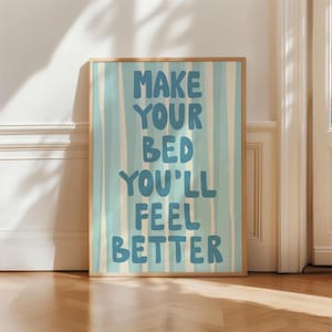 Könnte beinhalten: Ein gerahmter Kunstdruck mit dem Text "MAKE YOUR BED YOU'LL FEEL BETTER" in Blau, vor einem hellblauen und cremefarbenen gestreiften Hintergrund. Der Rahmen ist hellbraun. Das Kunstwerk wird auf einem Holzboden ausgestellt.