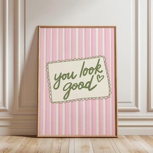 May include: A framed print with a pink and white striped background. The print features a cream-colored rectangle with the words "you look good" in green script, along with a small heart. The frame is a light brown color.