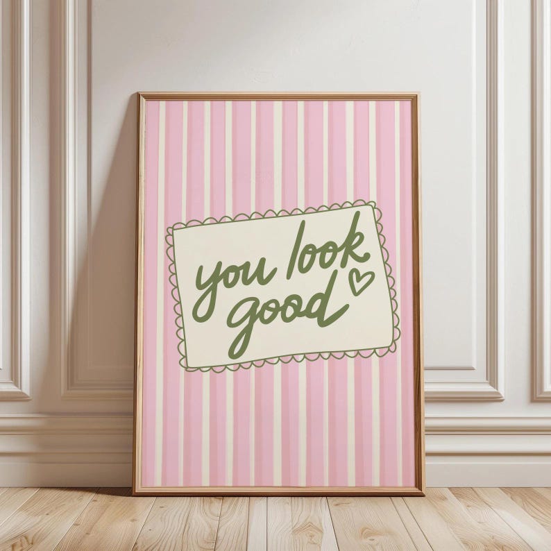 May include: A framed print with a pink and white striped background. The print features a cream-colored rectangle with the words "you look good" in green script, along with a small heart. The frame is a light brown color.