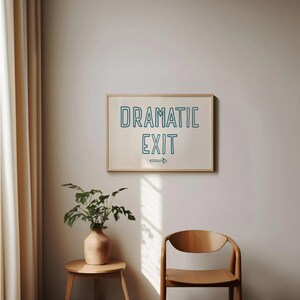 May include: A framed print with the text "Dramatic Exit" in blue outlined lettering with an arrow pointing to the right. The print is hanging on a white wall with a wooden frame. A potted plant and a wooden chair are in the foreground.