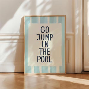 May include: A framed art print with the words "GO JUMP IN THE POOL" in navy blue on a cream background. The print is bordered by light blue vertical stripes and a wooden frame. The artwork is displayed on a wooden floor.