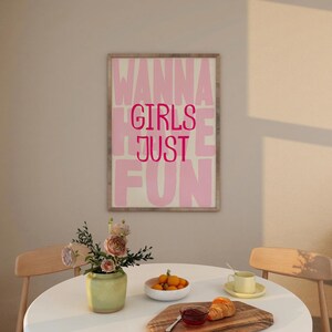 May include: A framed print with the words "WANNA GIRLS JUST FUN" in pink letters. The print hangs above a table set with a vase of flowers, a bowl of oranges, and a wooden board with a croissant and jam.