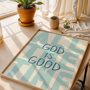 May include: A framed art print with the words "ALL GOD IS GOOD" in blue over a light blue and cream background. The artwork is displayed on a wooden table near a window with a potted plant and a beige cup.