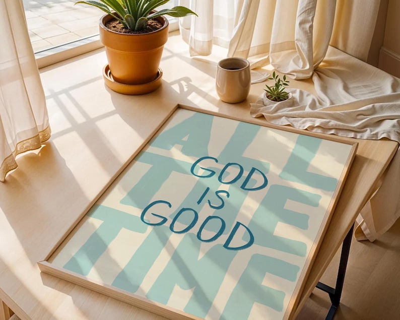 May include: A framed art print with the words "ALL GOD IS GOOD" in blue over a light blue and cream background. The artwork is displayed on a wooden table near a window with a potted plant and a beige cup.