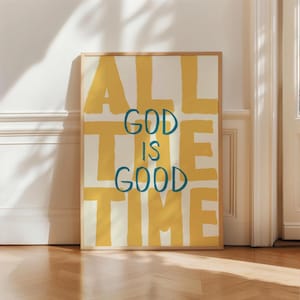 May include: A rectangular wall art print in a light wood frame featuring a cream background with large, bold, mustard yellow block letters spelling ALL THE TIME. Centered over the yellow text is the phrase GOD IS GOOD in dark blue script.