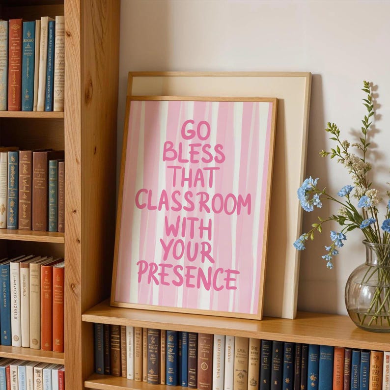 May include: A framed pink and white striped print with the text "GO BLESS THAT CLASSROOM WITH YOUR PRESENCE" in pink lettering. The print is displayed on a wooden shelf with a bookshelf in the background.