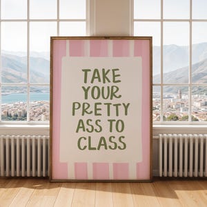 May include: A framed art print with the text "TAKE YOUR PRETTY ASS TO CLASS" in green lettering. The print has a pink and white striped border and is displayed in a room with a window overlooking a city and mountains.