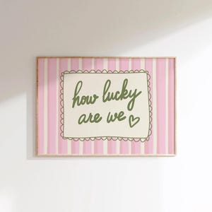 May include: A framed print with a pink and white striped background. The print features the phrase "how lucky are we" in green script within a scalloped-edged frame. A small heart is next to the word "we".