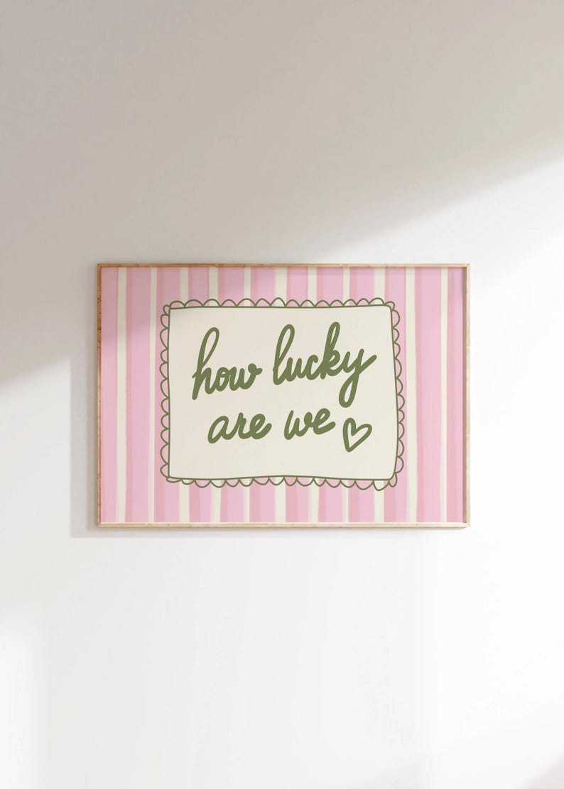 May include: A framed print with a pink and white striped background. The print features the phrase "how lucky are we" in green script within a scalloped-edged frame. A small heart is next to the word "we".