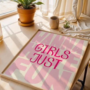 May include: A framed art print with the words "GIRLS JUST WANNA HAVE FUN" in pink lettering on a light pink and cream background. The print is displayed on a wooden table near a window with a potted plant.