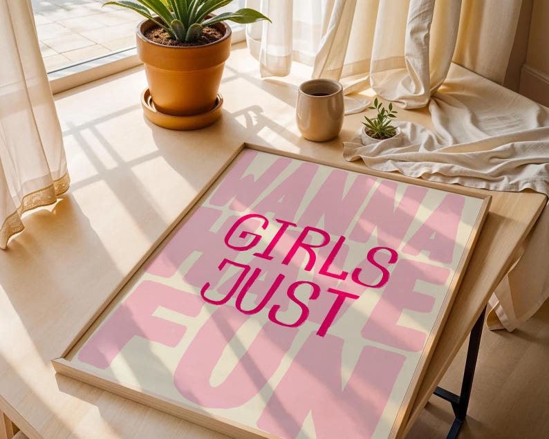 May include: A framed art print with the words "GIRLS JUST WANNA HAVE FUN" in pink lettering on a light pink and cream background. The print is displayed on a wooden table near a window with a potted plant.