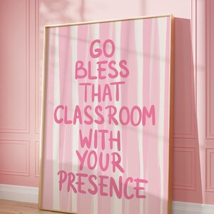 May include: A framed pink and white striped art print with the text "GO BLESS THAT CLASSROOM WITH YOUR PRESENCE" in pink lettering. The artwork is displayed against a pink wall and wooden floor.