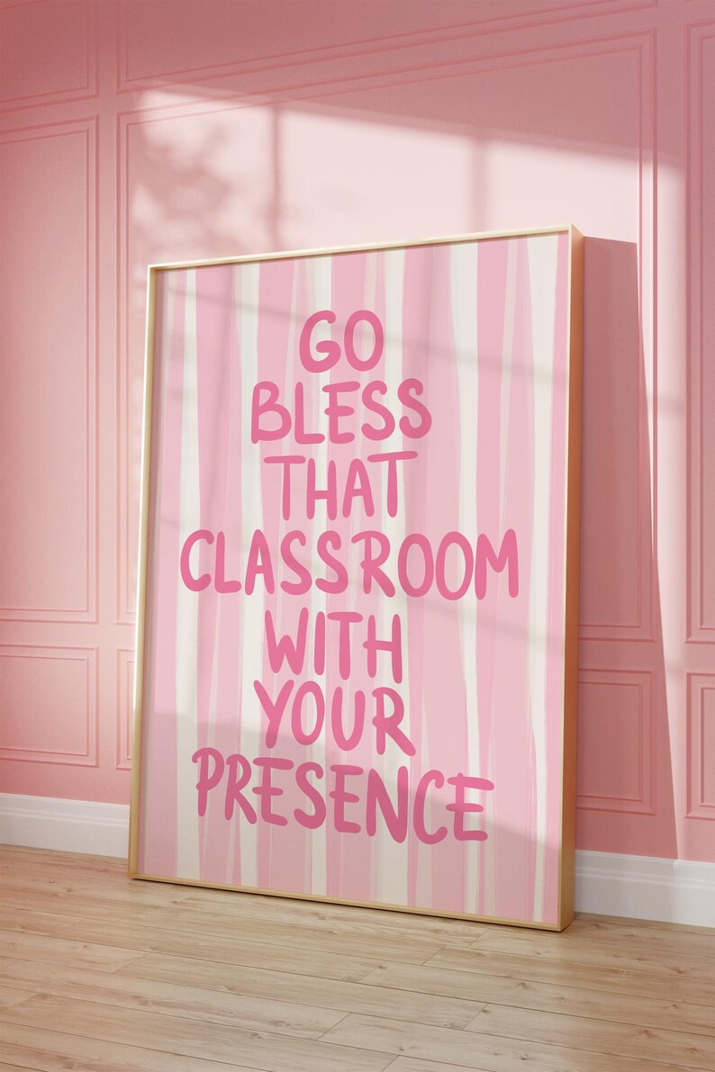 May include: A framed pink and white striped art print with the text "GO BLESS THAT CLASSROOM WITH YOUR PRESENCE" in pink lettering. The artwork is displayed against a pink wall and wooden floor.