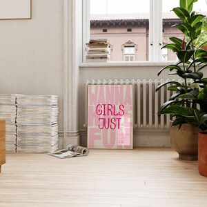 May include: A framed poster with the words "WANNA GIRLS JUST FUN" in pink and white lettering. The poster is leaning against a radiator in a room with a large window and potted plants. The floor is light wood.