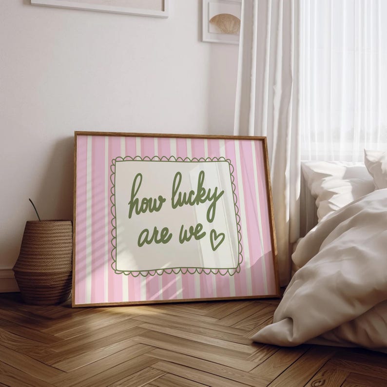 May include: A framed print with a pink and white striped background. The print features the phrase "how lucky are we" in green script, enclosed in a scalloped border. A small heart is next to the word "we". The frame is a light brown color.