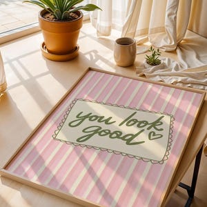 May include: A framed print with a pink and white striped background. The print features the phrase "you look good" in green script within a decorative border. The frame is a light wood color.