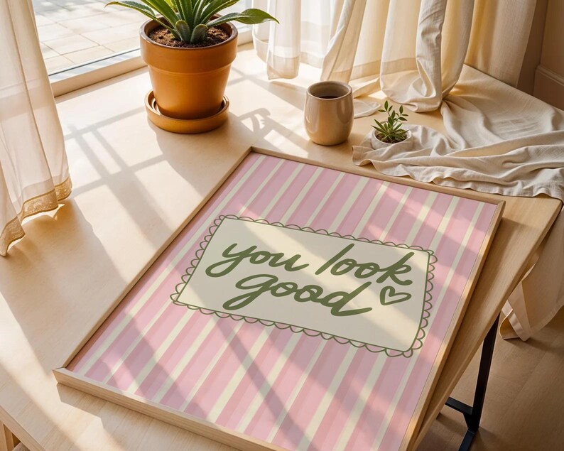 May include: A framed print with a pink and white striped background. The print features the phrase "you look good" in green script within a decorative border. The frame is a light wood color.