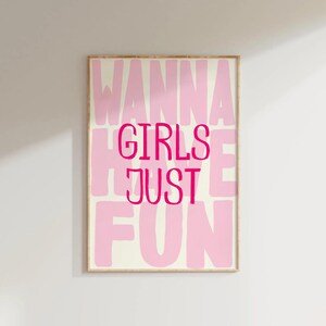 May include: A framed art print with the words "WANNA GIRLS JUST FUN" in pink lettering. The words are stacked vertically and horizontally on a cream-colored background. The frame is a light brown color.