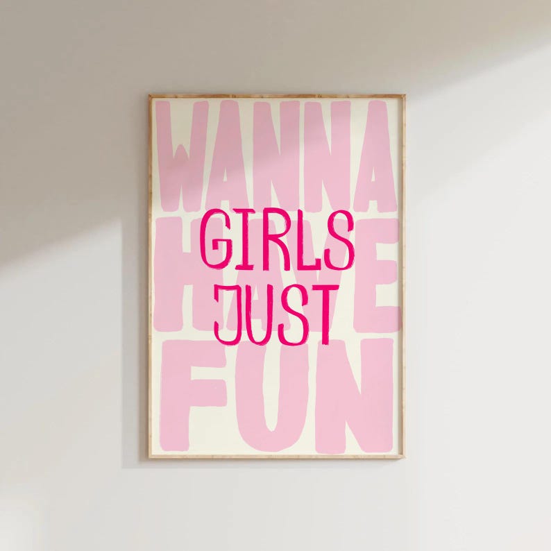 May include: A framed art print with the words "WANNA GIRLS JUST FUN" in pink lettering. The words are stacked vertically and horizontally on a cream-colored background. The frame is a light brown color.
