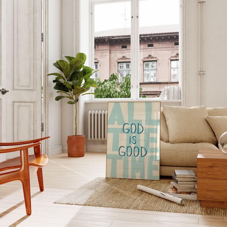 May include: A framed art print with the text "ALL GOD IS GOOD TIME" in light blue against a white background. The print is displayed in a living room setting with a beige sofa, wooden chair, and a potted plant.