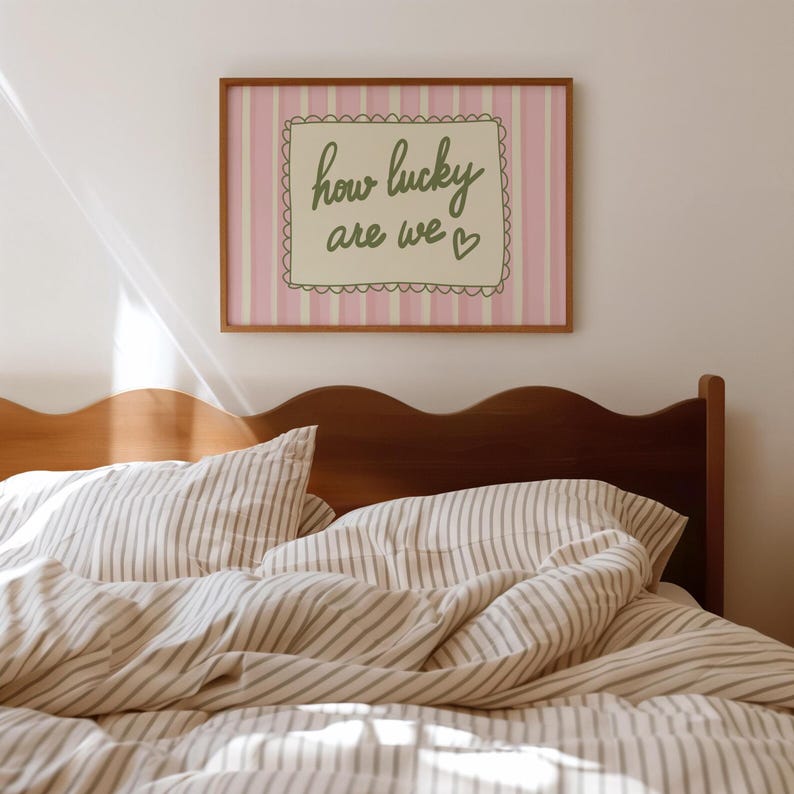 May include: A framed print with the text "how lucky are we" in a bedroom setting. The print has a pink and white striped background with a scalloped border. The bed has striped bedding and a wooden headboard.