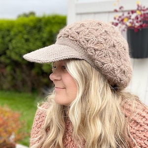 May include: A light brown crocheted newsboy cap featuring a textured star-like pattern across the crown and a solid, flat-stitched visor. The hat is crafted from a soft, fuzzy yarn and maintains a structured shape with a rounded top and brim.